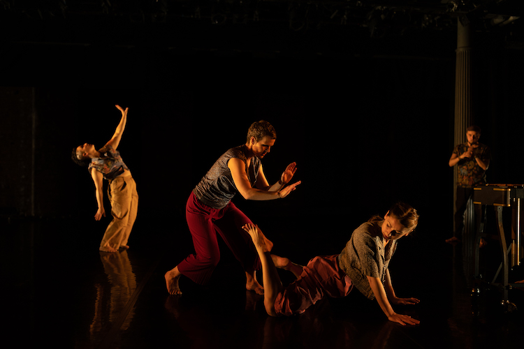 in dim light one dancer in loose fitting pants and a midriff top stands in profile while lifting one arm to the ceiling, in front of her are two other dancers in grey tops of varied sleeve length and dark pants,one dancer attempts to pat the back of another dancer whose lower body lays on the floor, while her upper body arches and twists supported by her arms, in the very back is a musician playing a flute 